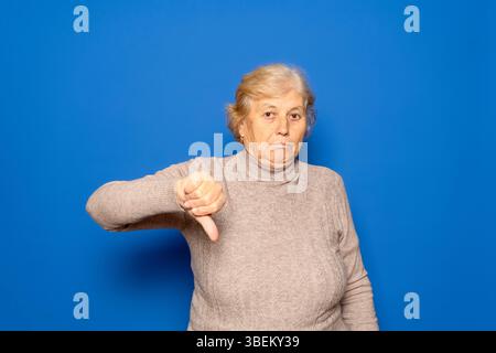 Weiße Frau in ihren 70ern, die einen dunkelbeigen Pullover mit dem Daumen nach unten trug, als Zeichen von Ekel und Missbilligung, isoliert auf blauem Studiohintergrund. Stockfoto