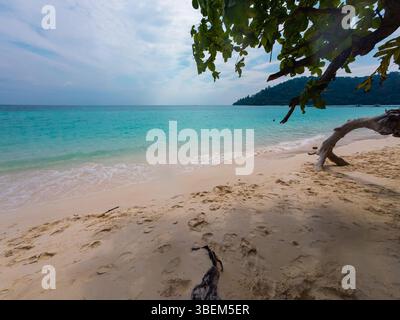 Sanfte Wellen schlagen gegen die Sandküste, während sich türkisfarbenes Wasser zum Horizont ausbreitet. Üppiges Grün umrahmt diesen ruhigen, einladenden Küstenort Stockfoto