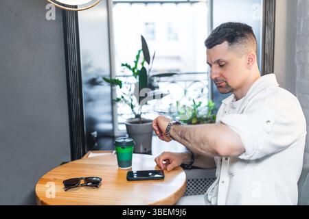 Ein fokussierter Geschäftsmann in weißer Kleidung schaut seine Armbanduhr in einem stilvollen, modernen Café. Ein Smartphone liegt in der Nähe auf dem Tisch. Stockfoto