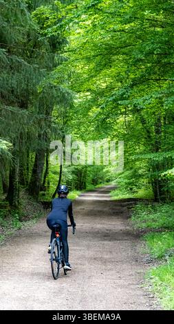 Ein Radfahrer in schwarzer Kleidung fährt auf einem ruhigen Waldweg, der im Frühjahr von üppig grünen Bäumen umgeben ist. Das Bild fängt die Ruhe und Schönheit von ein Stockfoto