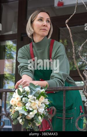 Floristin in grüner Schürze mit einem Strauß weißer und pfirsichfarbener Rosen vor einem Blumenladen. Blumendesign Stockfoto