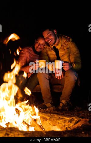 Eine gemütliche Szene am Lagerfeuer bei Nacht, die Wärme hervorhebt. Zwei Personen genießen heiße Getränke im Feuer glühen und tragen Jacken gegen die Kälte im Fo Stockfoto