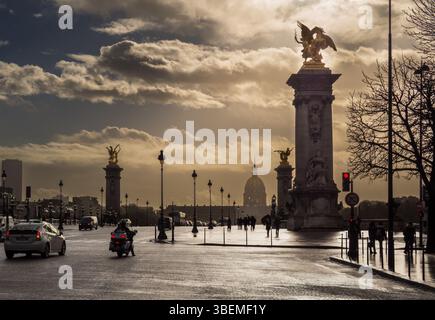 Wolken sammeln sich über der berühmten Alexandre III Brücke, während Fahrzeuge und Menschen durch die belebten Straßen von Paris fahren. Frankreich Stockfoto