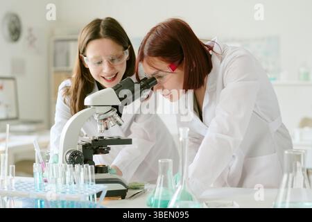 Zwei junge Wissenschaftlerinnen arbeiten im Labor zusammen und analysieren Proben mit Mikroskop, während sie Schutzbrillen tragen, umgeben von Laborgeräten Stockfoto
