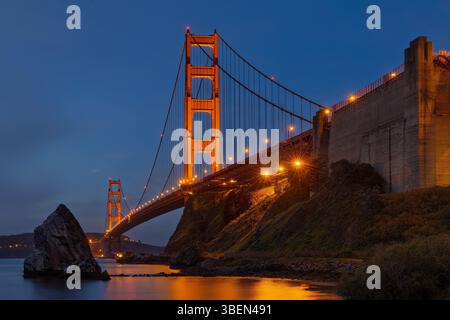 Die majestätische Golden Gate Hängebrücke erstreckt sich über ein ruhiges Gewässer, beleuchtet von warmen, leuchtenden Lichtern vor einem tiefblauen Abendhimmel. Stockfoto