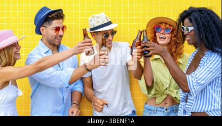 Fünf fröhliche junge Freunde in Sommerkleidung und Hüten toasten mit Bierflaschen und feiern eine Party vor einer leuchtend gelben Wand Stockfoto