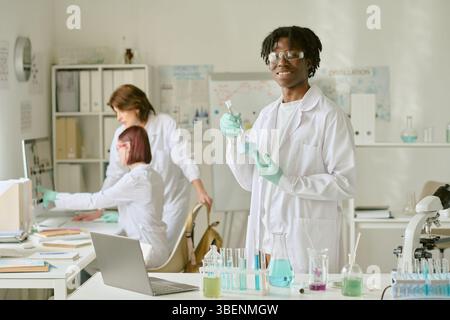 Student arbeitet mit Laborgeräten, führt Experimente durch. Zwei Kollegen, die sich mit der Forschung im Hintergrund beschäftigen. Laborumgebung mit wissenschaftlichen Werkzeugen Stockfoto