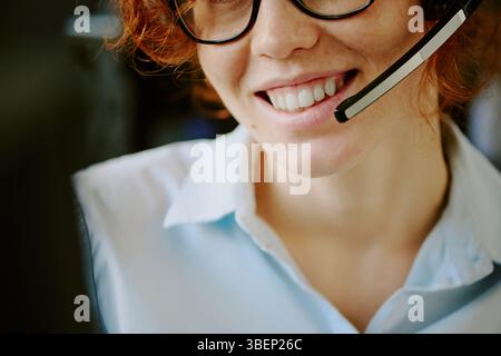 Lächelnde Frau mit lockigem rotem Haar und Brille, die Headset und Hemd trägt, im Büro arbeitet, Gespräche führt Stockfoto