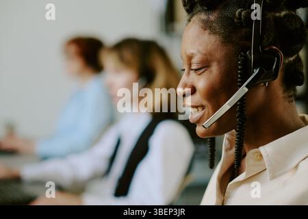 Afroamerikanerin lächelt, während sie das Headset benutzt, im Profil schaut. Andere Kundendienstmitarbeiter, die im Hintergrund arbeiten Stockfoto
