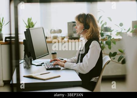 Frau mit Headset und Tastatur, während sie in einem modernen Büro mit Holzschreibtisch und großem Monitor arbeitet. Stockfoto