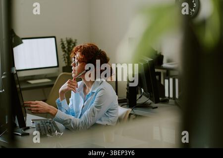 Frau, die am Schreibtisch in einem modernen Büro sitzt, Headset trägt und am Computerbildschirm Gespräche führt, mit Büroausstattung im Hintergrund Stockfoto