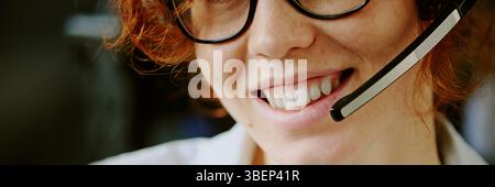 Porträt einer jungen Frau mit lockigem Haar, die beim Tragen von Headset und Brille lächelt. Glatte Haut und helle Zähne sichtbar für eine warme, freundliche Atmosphäre Stockfoto