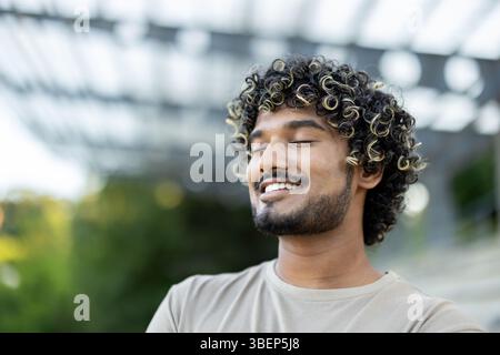 Ein fröhlicher Mann mit lockigen Haaren und Bart schließt die Augen und lächelt voller Zufriedenheit. Stockfoto