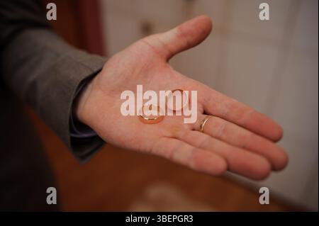 Eine Nahaufnahme zeigt die Hand eines Mannes, der zwei einfache goldene Eheringe hält, die Ehe, Liebe und Bindung symbolisieren, gegen ein verschwommenes, warmes ich Stockfoto