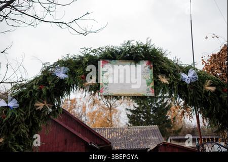 Ein rustikaler Hochzeitsbogen mit immergrünen Zweigen und Blumenarrangements steht an einem bewölkten Tag vor einem traditionellen roten Holzgebäude. Stockfoto