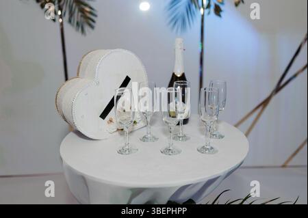 Eleganter Champagner auf einem weißen runden Tisch mit einer herzförmigen Box, einer Flasche Sekt und vier Gläsern, beleuchtet in einem sanft beleuchteten Raum mit wi Stockfoto