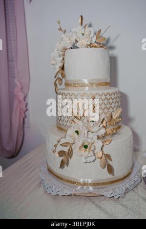 Eleganter dreistöckiger Hochzeitstorte mit raffinierten Details aus weißer Spitze, goldenen Akzenten und Blumendekorationen vor einem weichen rosafarbenen Hintergrund, der einen feiert Stockfoto