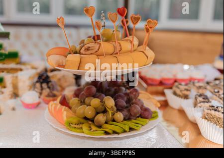 Eine gestufte Dessertplatte mit verschiedenen Früchten, einschließlich Bananen, Trauben, Kiwi und Käsewürfeln, dekoriert mit herzförmigen Zahnstochern, Presente Stockfoto