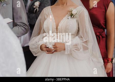 Eine strahlende Braut in einem atemberaubenden langärmeligen Spitzenkleid steht während einer religiösen Zeremonie mit einem Bräutigam und hält Kerzen in einer warm beleuchteten Kirche Stockfoto