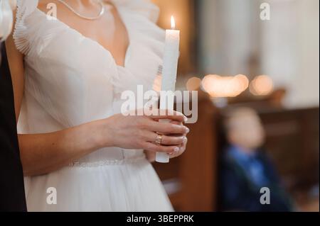Eine strahlende Braut in einem weißen Rüschenkleid hält während einer Hochzeitszeremonie in einer Kirche eine Kerze, die Glauben und Einheit mit einem Unschärfen symbolisiert Stockfoto