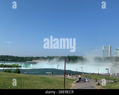 Touristen genießen die Aussicht auf die Niagarafälle und den Nebel, der von den Horseshoe Falls an einem klaren Sommertag von der US-Seite mit Kanadas Skyline aufsteigt. Stockfoto