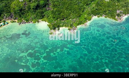 Blick aus der Vogelperspektive auf klares türkisfarbenes Wasser, das sanft auf eine üppig grüne Küste mit felsigen Ausbissen trifft. Praslin, Seychellen. Stockfoto