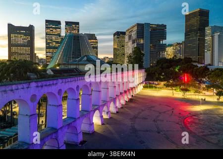 Lapa Aquädukt, beleuchtet mit violetten und rosa Farben am Abend mit hohen Bürogebäuden in der Innenstadt von Rio de Janeiro Stockfoto
