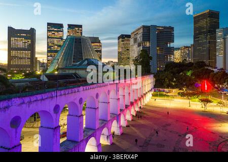 Lapa Aquädukt, beleuchtet mit violetten und rosa Farben am Abend mit hohen Bürogebäuden in der Innenstadt von Rio de Janeiro Stockfoto