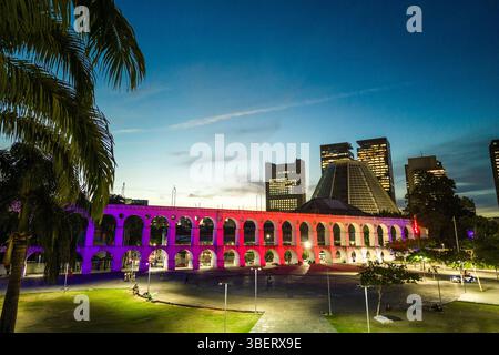 Lapa Aquädukt, beleuchtet mit violetten und rosa Farben am Abend mit hohen Bürogebäuden in der Innenstadt von Rio de Janeiro Stockfoto
