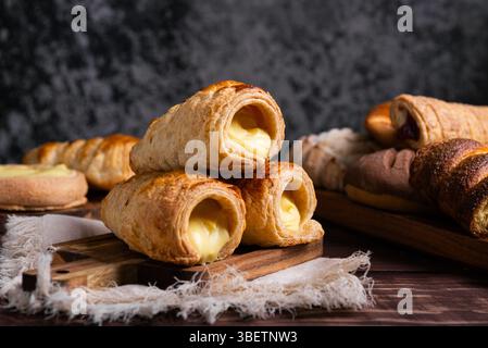 Eine verlockende Auswahl an frisch gebackenem Gebäck, mit cremigem Pudding gefülltem Blätterteig auf einem rustikalen Holzbrett. Stockfoto