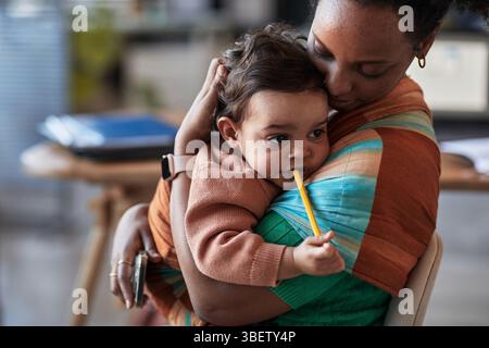 Afroamerikanerin hält Kind in den Armen, während Kind Bleistift hält. Ungezwungenes Ambiente im Innenbereich mit Möbeln und Haushaltsgegenständen Stockfoto