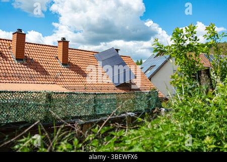 Solarpaneele auf rot gekachelten Dächern von Häusern, umgeben von üppigem Grün und blauem Himmel. Umweltfreundliche Energielösungen im ländlichen Raum Stockfoto