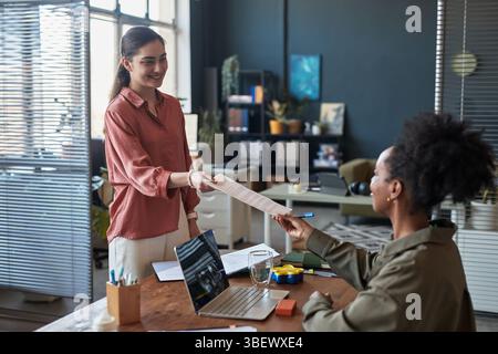 Zwei Frauen teilen sich Dokumente in einer modernen Büroumgebung, die kollegiale Interaktion und Kommunikation zeigen. Die Szene beinhaltet einen Schreibtisch mit Laptop und m Stockfoto