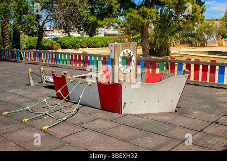 Niedrige Spielstruktur im Stil eines Piratenschiffes mit Seilnetzen in einem sonnigen Park. Umgeben von bunten Holzzaun, Bäumen und Palmen, Stockfoto
