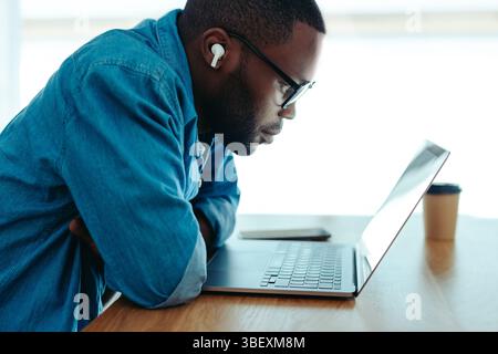 Ein afrikanischer Mann, der sich auf E-Learning konzentriert, sitzt mit einem Laptop an einem Schreibtisch und trägt kabellose Ohrhörer. Die Umgebung ist ruhig mit natürlichem Licht, Capturin Stockfoto