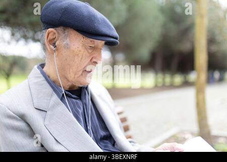Ein älterer Mann hört Musik mit Ohrhörern und liest Bücher im Park Stockfoto