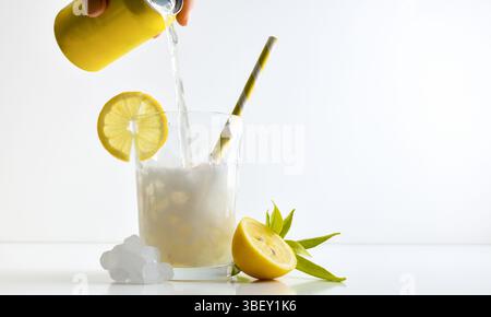 Erfrischendes Zitronengetränk mit Eis in einer Glasschale, dekoriert mit einem Strohhalm und einer Scheibe Obst auf einem weißen Tisch mit Obst, Eis und Blättern mit einem weißen IS Stockfoto