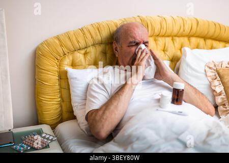 Kranker und erschöpfter älterer Mann, der Grippe hat und in Gewebe niest, während er auf dem Bett liegt, umgeben von Medikamenten zu Hause. Stockfoto