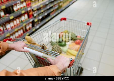 Person schiebt den Einkaufswagen mit frischen Produkten, einschließlich Obst wie Ananas und Bananen, in den Supermarktgang gefüllt mit verschiedenen Lebensmitteln Stockfoto