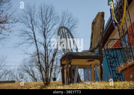 Zwei nicht passende Stühle, einer alt und verwittert, sitzen im Frühjahr vor einem bunten Haus auf einem grasbewachsenen Rasen. Blattlose Bäume und ein klarer blauer Himmel Stockfoto