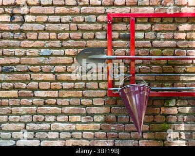 Brandbekämpfungsset an Ziegelwand mit rotem Metallrahmen, konischem Eimer, Schaufel und Haken, Sicherheitsausrüstung für den Einsatz in Notfällen, am Arbeitsplatz und in der öffentlichen Sicherheit. Stockfoto