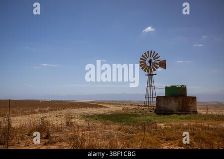 Eine alte Windmühle und ein grüner Wassertank stehen auf einem trockenen Feld im ländlichen Südafrika, mit Bergkämmen in der Ferne unter blauem Himmel. Stockfoto