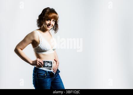 Eine junge Frau in einem weißen Spitzenbralette und Jeans lächelt stolz, während sie ein Ultraschallbild über ihrem Bauch zeigt, das Freude und Begeisterung symbolisiert Stockfoto