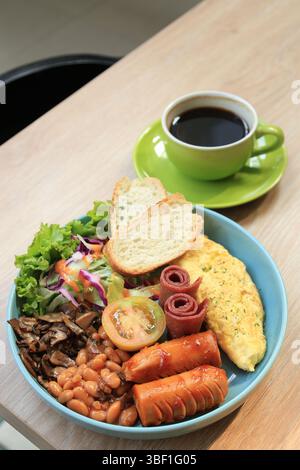 Englisches Frühstück mit Bohnen, Speck, Eieromelette, Salat, Brot, und Würstchen. Serviert mit schwarzem Kaffee, Morning Cafe Restaurant Menü Stockfoto