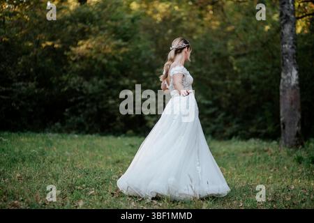 Das Bild zeigt eine Braut in einem eleganten weißen Hochzeitskleid in einer ruhigen Waldumgebung. Das Kleid ist mit zarten Spitzendetails versehen Stockfoto
