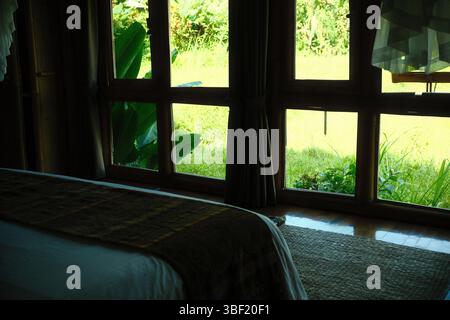 Ein Blick vom Inneren eines Bungalow-Schlafzimmers, mit Blick auf die üppige grüne Vegetation durch große Holzfenster Stockfoto