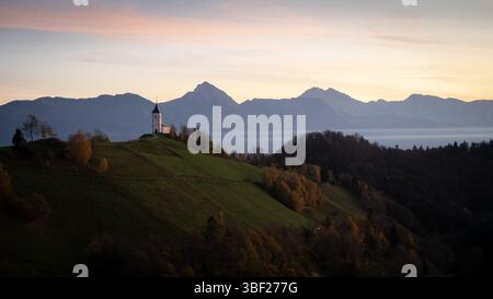 Landschaft mit einer kleinen Kirche auf einem Hügel und Bergen im Hintergrund während des Sonnenaufgangs, Slowenien Stockfoto