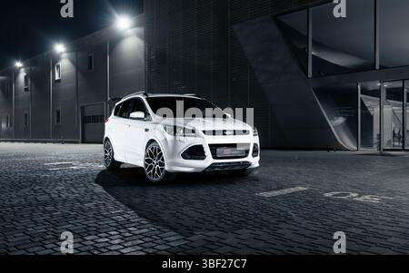 Weißer Ford Kuga SUV in der Nacht auf einem gepflasterten Parkplatz - Vorderansicht. Modernes Gebäude mit leuchtenden Projektoren im Dunkeln. Stockfoto