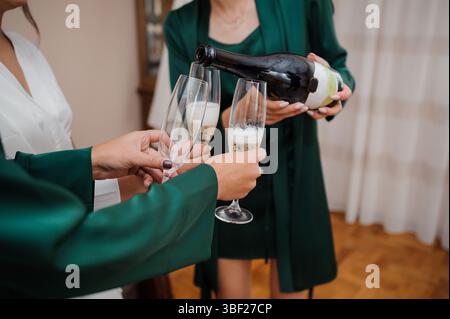 Eine gemütliche Szene mit zwei Frauen, wahrscheinlich Brautjungfern, die sich auf eine Hochzeit vorbereiten, eine mit Champagner in Gläser gießt, während sie sie hält, um ein f Stockfoto