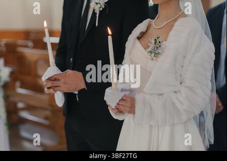 Eine Nahaufnahme einer orthodoxen Hochzeitszeremonie mit einer Braut und einem Bräutigam, die beleuchtete Kerzen halten, die Einheit und den Glauben in einem wunderschönen Kirchenset symbolisieren Stockfoto